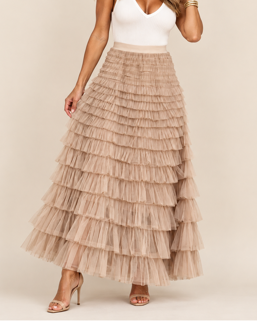 A woman modeling a long, tiered tulle maxi skirt in a light taupe-beige color, paired with a white ribbed tank top and nude high-heeled sandals, against a solid beige studio background.