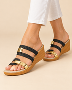 A pair of women's wedge sandals with black glittery straps and shiny gold metallic accents, shown on a model wearing white pants against a beige studio background.