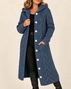 A woman wearing a long, blue cable-knit hooded cardigan with large white buttons, styled over a black top and black pants, against a plain beige background.