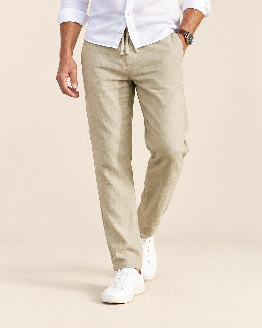 A man wearing beige drawstring trousers, a white button-down shirt, and white sneakers, shown from the waist down against a light beige studio background.