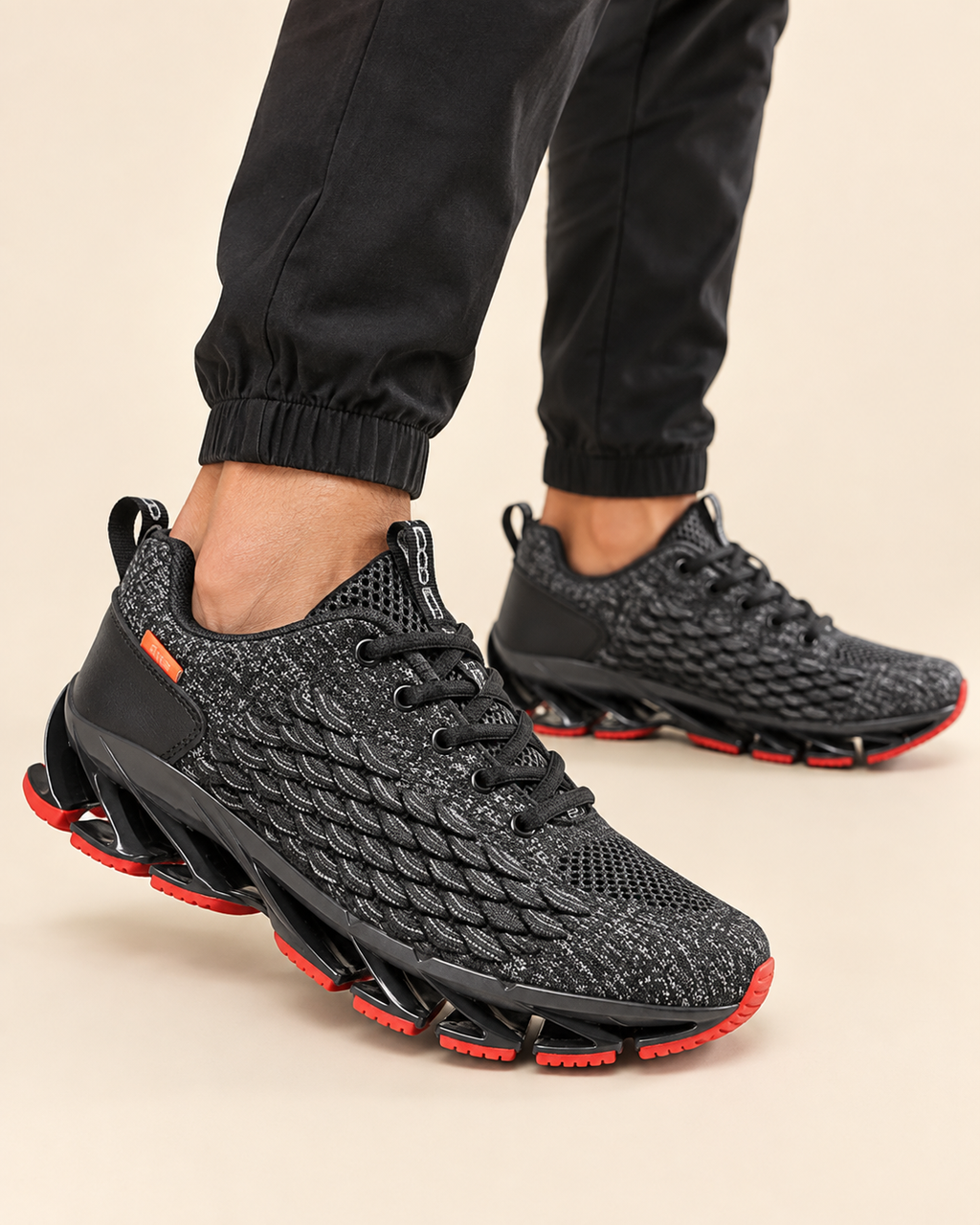 A pair of black textured athletic sneakers with bright red outsole accents, shown on a model wearing black joggers against a light beige background.