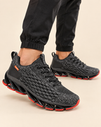 A pair of black textured athletic sneakers with bright red outsole accents, shown on a model wearing black joggers against a light beige background.