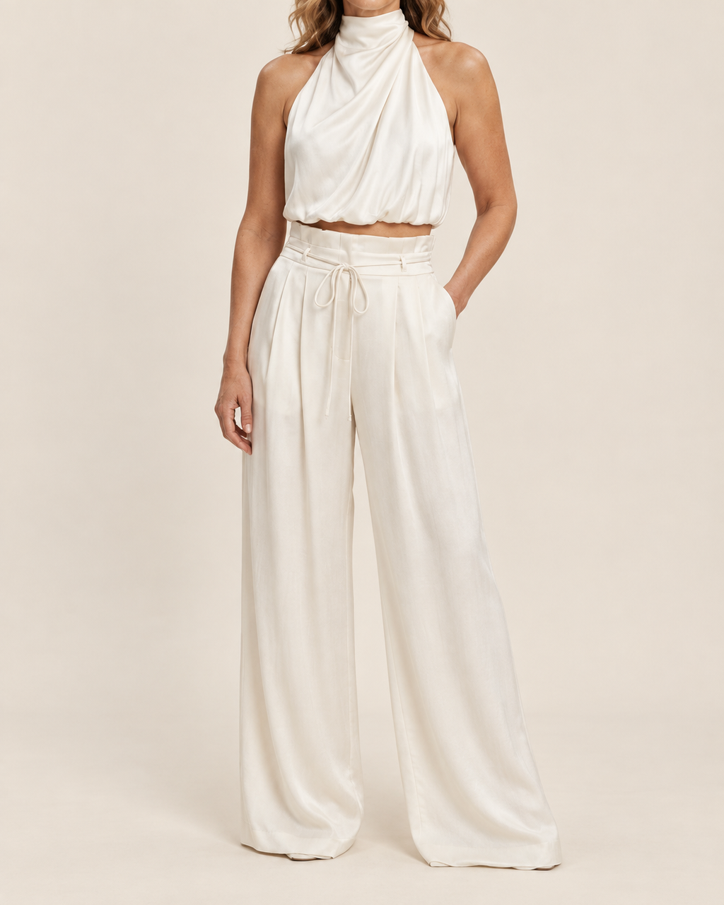 A model wearing a matching two-piece outfit consisting of a high-neck draped halter crop top and high-waisted wide-leg trousers in a warm off-white or cream color, set against a solid light beige studio background.