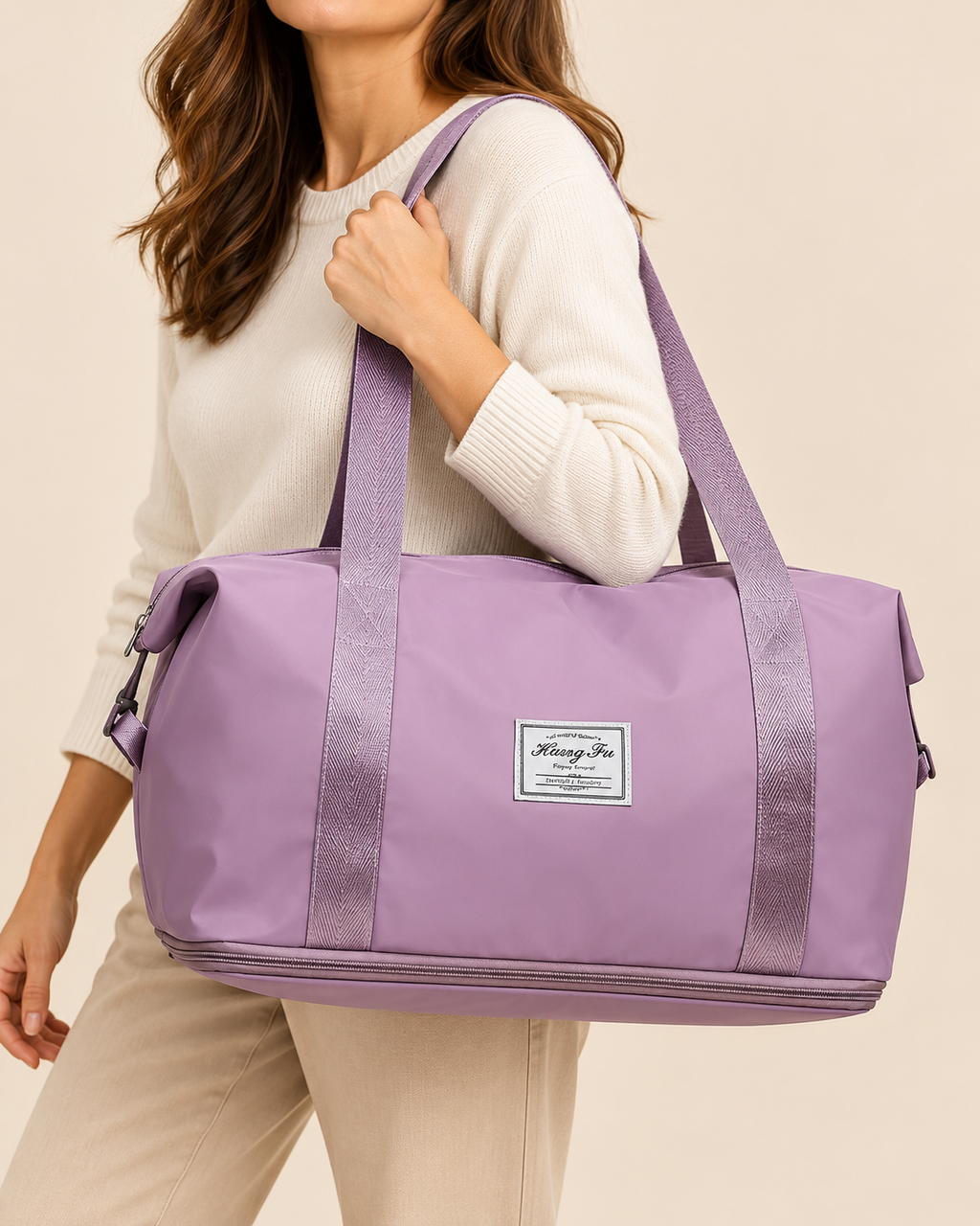 A woman wearing a cream knit sweater and beige pants carries a large lavender purple duffel bag with matching shoulder straps. The bag has a white rectangular brand patch on the front that reads 'Fancy Fun'.