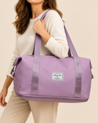 A woman wearing a cream knit sweater and beige pants carries a large lavender purple duffel bag with matching shoulder straps. The bag has a white rectangular brand patch on the front that reads 'Fancy Fun'.