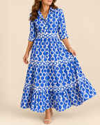 A woman wearing a blue and white chain-link print maxi dress with three-quarter sleeves and a tiered skirt, standing against a solid beige studio background.