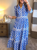 A woman wearing a blue and white chain-link patterned maxi dress, standing in a bedroom setting next to a wooden nightstand and holding a metallic gold handbag.