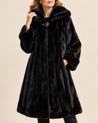 A woman wearing a luxurious black faux fur coat with a wide collar and a decorative silver-toned brooch at the neckline, set against a solid beige background.