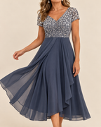 A woman wearing a navy blue midi dress with a sequined silver bodice and sheer layered skirt, posed against a solid beige background.