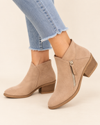 A pair of beige suede ankle boots with a side zipper and stacked block heel, shown on a model wearing light blue frayed-hem jeans against a solid cream background.