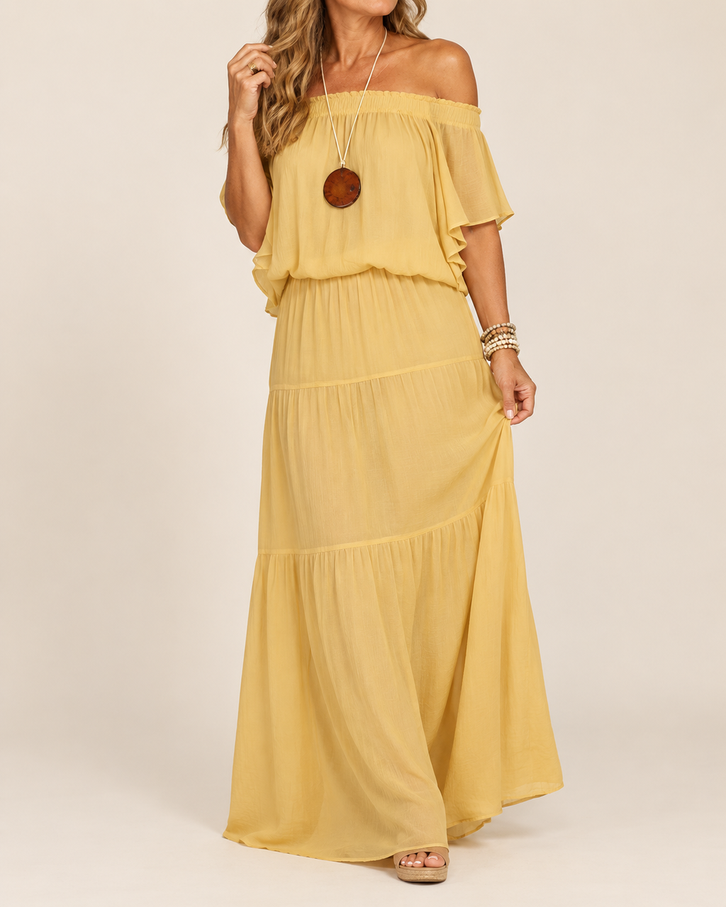 A woman modeling a flowing off-the-shoulder maxi dress in a soft mustard yellow color, paired with a long brown pendant necklace and bracelets, against a plain beige studio background.
