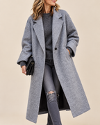 A woman wearing a long, double-breasted gray wool-blend coat over a gray sweater and distressed gray jeans, holding a black clutch against a solid beige background.