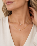A close-up of a woman wearing a delicate double-layered gold necklace with two small white freshwater pearls, styled with a cream-colored top.
