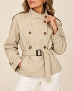 A woman wearing a beige double-breasted trench coat with a matching waist belt and white pants, shown from the chin down against a plain light beige background.