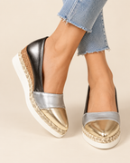 A pair of women's platform wedge espadrilles in metallic silver and gold, styled with frayed blue jeans on a light beige background.