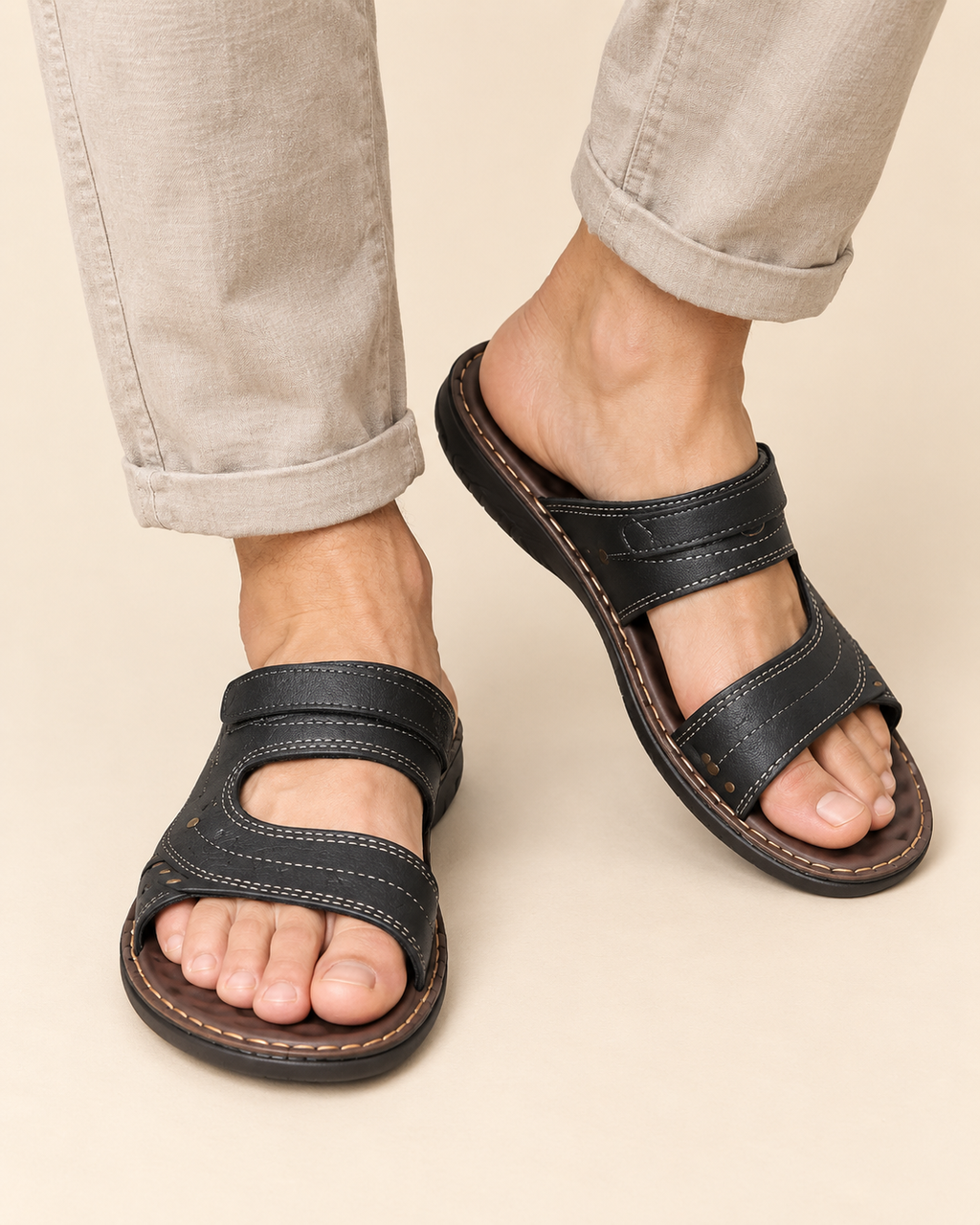 A pair of men's black leather slide sandals with double straps and contrast stitching, shown on a model wearing light beige pants against a solid beige background.