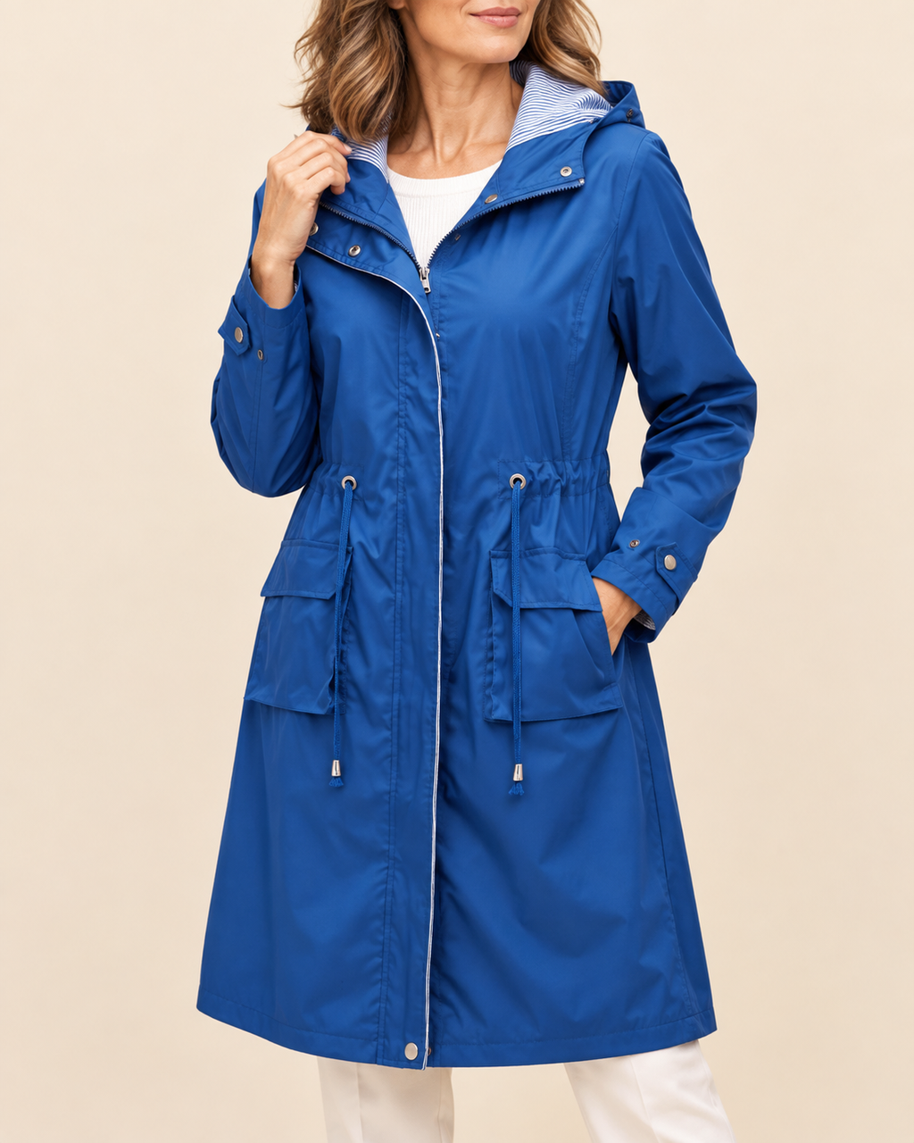 A woman wearing a bright blue hooded raincoat over a white top and white pants, standing against a plain beige studio background.