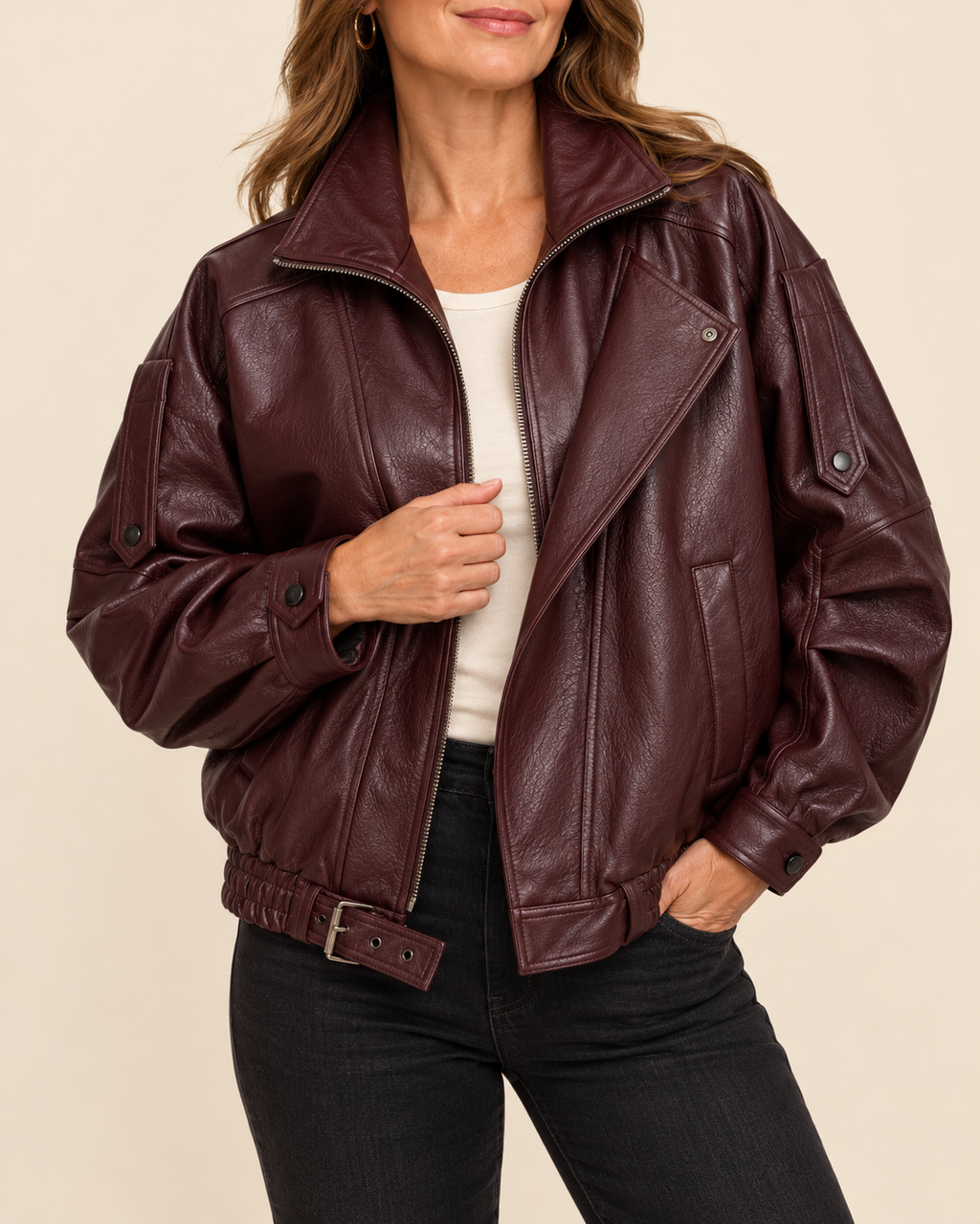 A woman wearing a dark burgundy faux-leather moto jacket with an asymmetrical zipper, belted hem, and ribbed cuffs, styled with a white top and black jeans against a solid beige background.