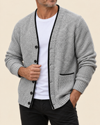 A man wearing a gray textured knit cardigan with black trim over a white t-shirt and black pants, shown from the chin down against a solid beige background.