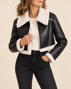 A woman wearing a black faux-leather moto jacket with a thick white sherpa lining, styled over a cream ribbed top and black jeans against a solid beige background.