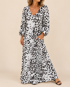 A woman wearing a long white maxi dress with a black leopard print pattern, featuring balloon sleeves and a V-neckline, standing against a solid beige background.