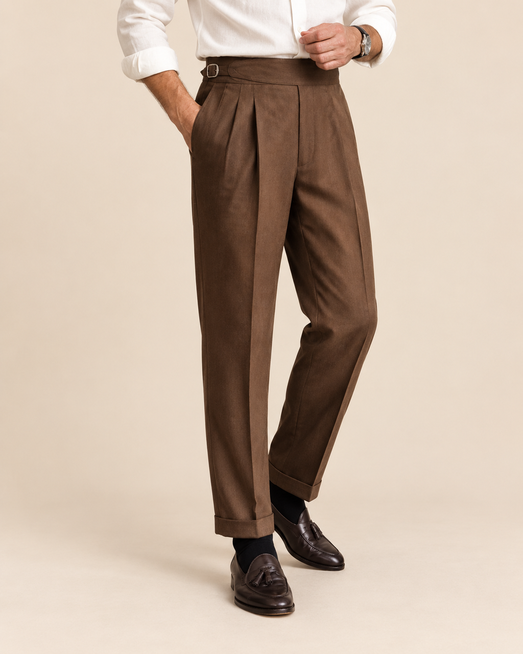 A man wearing brown pleated trousers, a white dress shirt, and dark brown leather loafers, standing against a solid beige studio background.