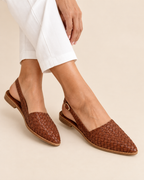 A person wearing brown woven slingback flats with pointed toes and white cropped pants, shown against a plain beige studio background.