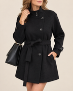 A woman wearing a black belted double-breasted coat with a mock neck, paired with a black handbag, against a solid beige background.