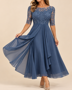 A woman modeling a dusty blue A-line midi dress with a lace bodice and sheer lace sleeves, holding out the flowing chiffon skirt against a solid beige background.