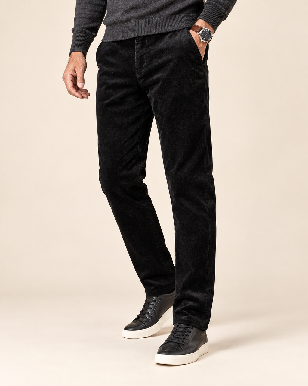 A person wearing slim black corduroy trousers, a dark gray knit sweater, and black low-top sneakers with white soles, standing against a plain beige studio background.