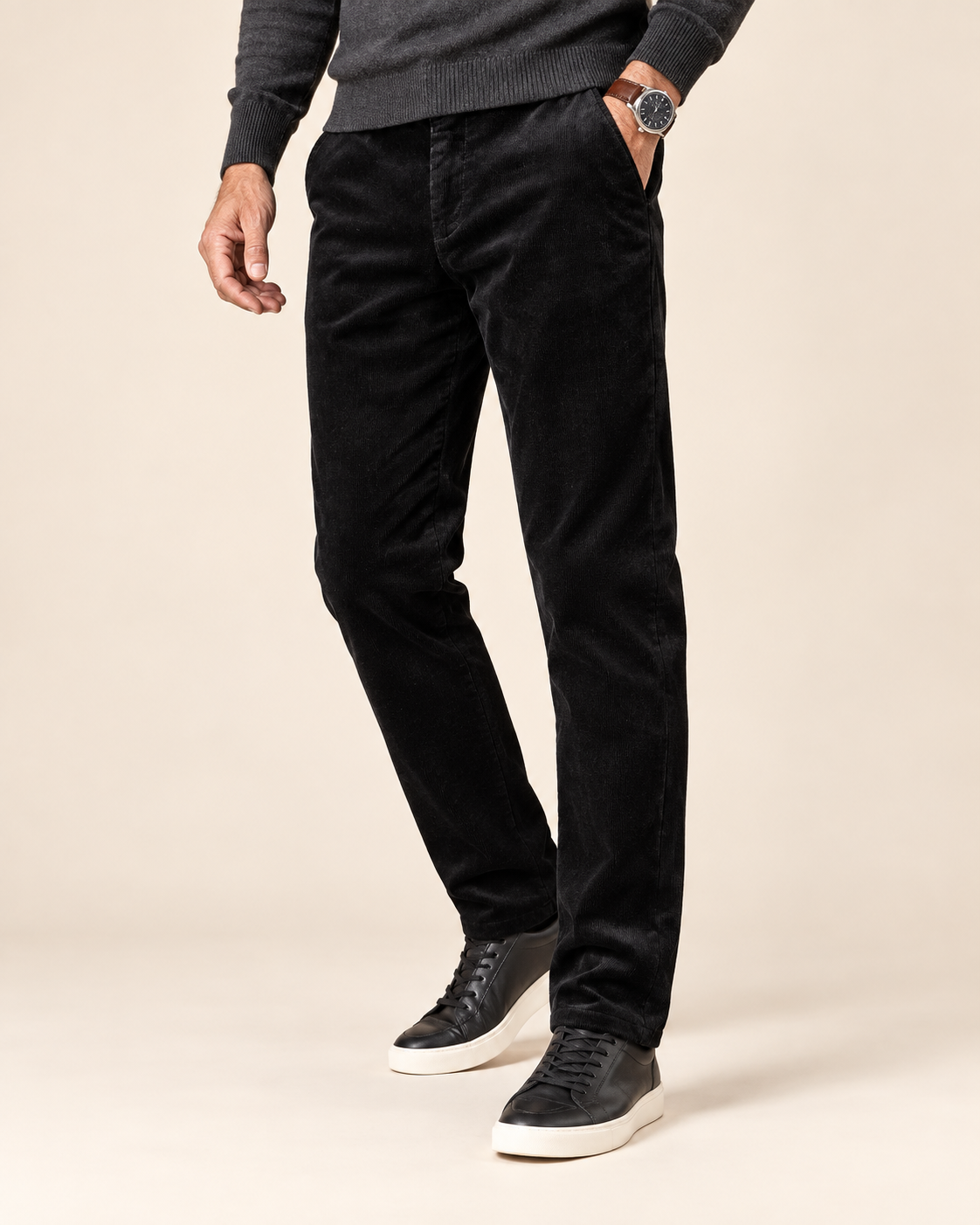 A person wearing slim black corduroy trousers, a dark gray knit sweater, and black low-top sneakers with white soles, standing against a plain beige studio background.