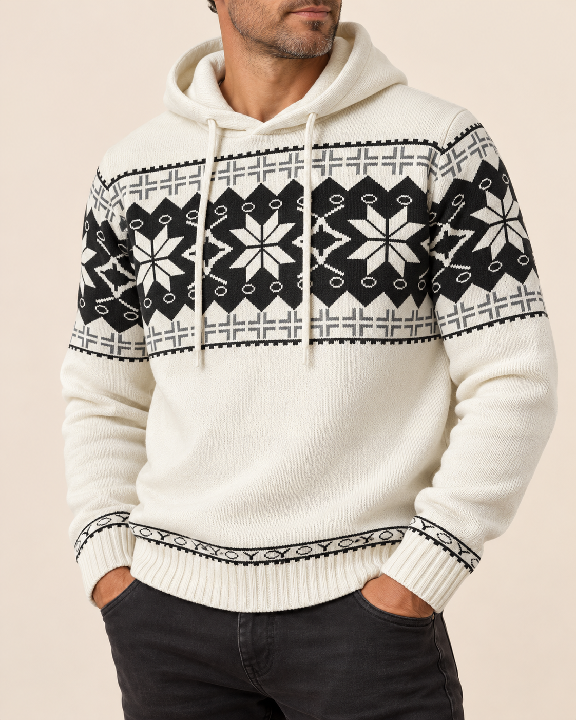 A man wearing a cream-colored knit hoodie with a black and white Fair Isle pattern across the chest, shoulders, and cuffs, paired with dark gray jeans against a solid beige background.