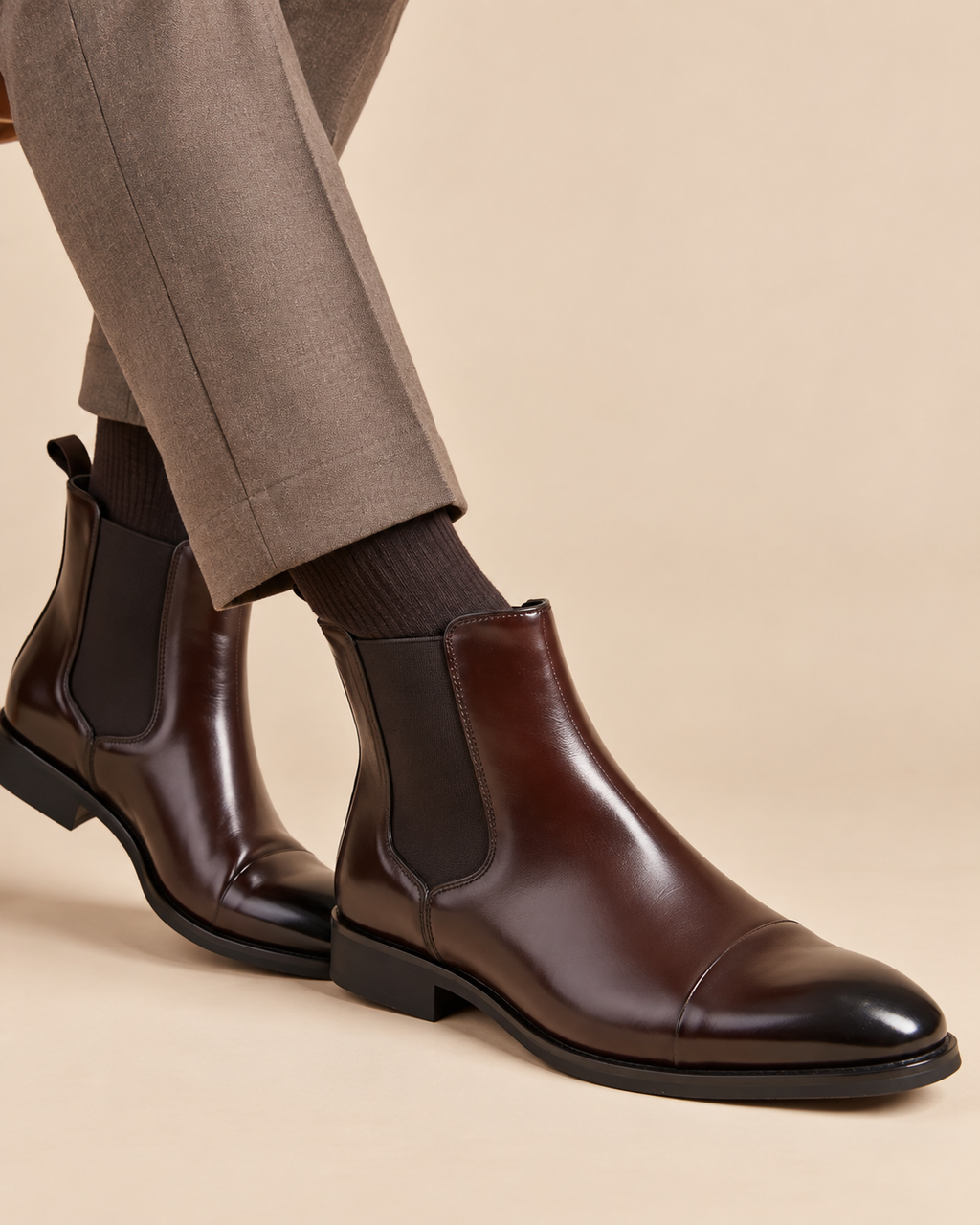 A pair of dark brown leather Chelsea boots with black elastic side panels, worn with dark socks and taupe trousers against a solid beige background.