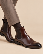 A pair of dark brown leather Chelsea boots with black elastic side panels, worn with dark socks and taupe trousers against a solid beige background.