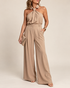 A woman models a beige halter-neck jumpsuit with a tie detail at the collar and wide-leg pants, holding a matching clutch against a plain beige studio background.