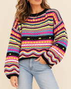 A woman wearing a vibrant, multicolored knit sweater with horizontal wavy stripes in pink, yellow, blue, orange, and black, paired with light-wash blue jeans against a solid beige background.