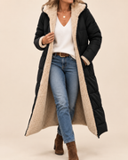 A woman models a long black puffer coat with a thick beige sherpa lining, styled with a white V-neck top, blue jeans, and brown ankle boots against a solid beige background.