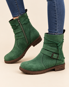 A pair of green suede ankle boots with side zippers, buckle straps, and wraparound laces, shown on a model wearing blue jeans against a light beige background.