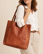 A woman wearing a white linen shirt and beige trousers carries a large cognac brown leather tote bag over her shoulder against a warm beige studio background.