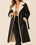 A woman wearing a long black faux-leather coat with a thick cream sherpa lining and collar, styled over a beige top and black pants against a plain beige background.