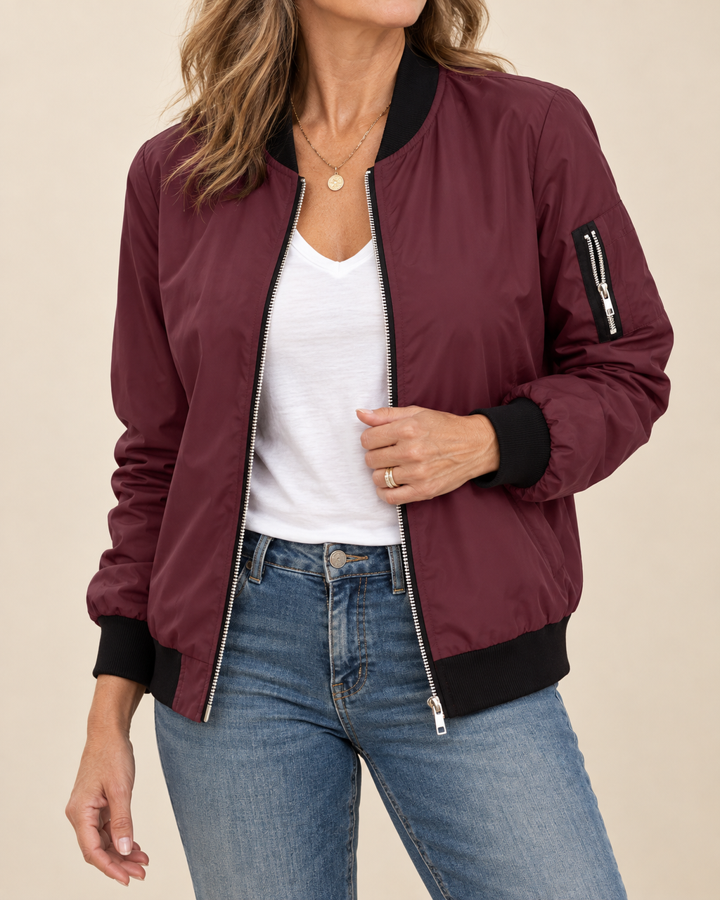 A woman wearing a burgundy bomber jacket with black ribbed collar, cuffs, and hem, styled over a white top and blue jeans against a solid beige background.