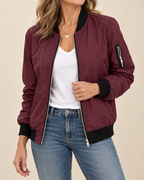 A woman wearing a burgundy bomber jacket with black ribbed collar, cuffs, and hem, styled over a white top and blue jeans against a solid beige background.
