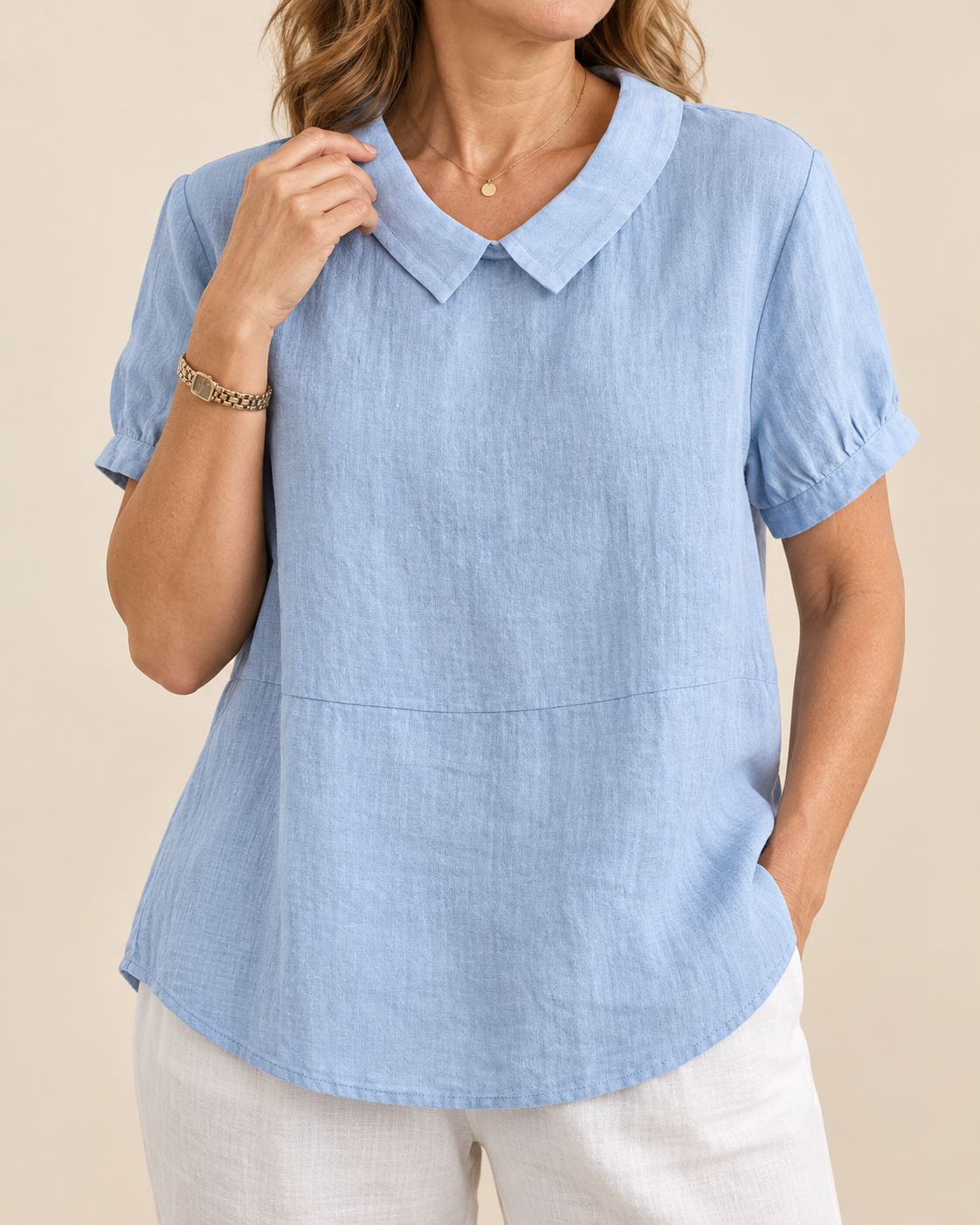 A woman wearing a light blue short-sleeve collared blouse with a curved hem and white pants, photographed against a plain beige studio background.