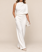 A woman modeling a white sleeveless mock-neck top and matching wide-leg trousers, styled with beige pointed-toe heels and a small white clutch against a warm beige studio background.