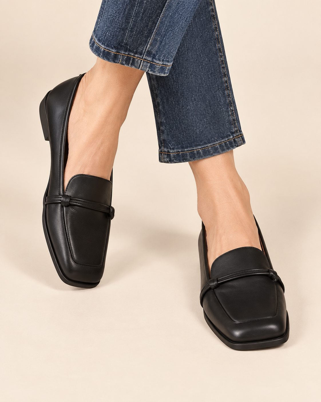 A pair of black leather square-toe loafers worn with blue jeans, shown from the side against a solid beige background.