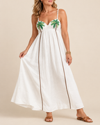 A woman modeling a white maxi dress with green palm tree embroidery on the bust and thin brown side stripes, photographed against a plain beige studio background.