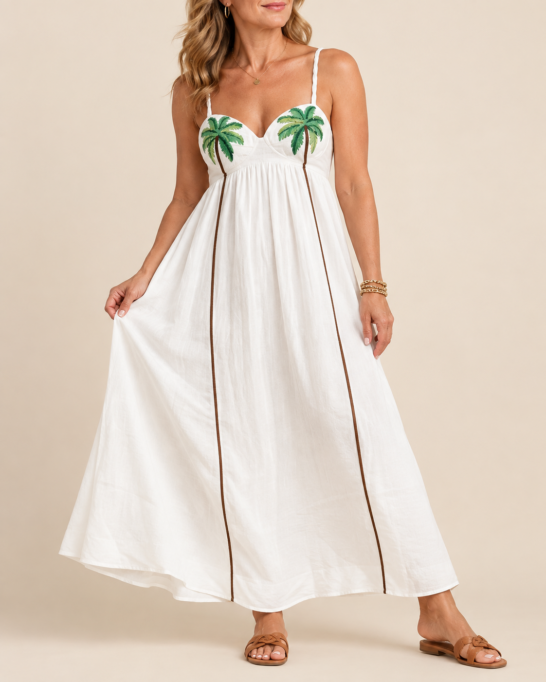A woman modeling a white maxi dress with green palm tree embroidery on the bust and thin brown side stripes, photographed against a plain beige studio background.